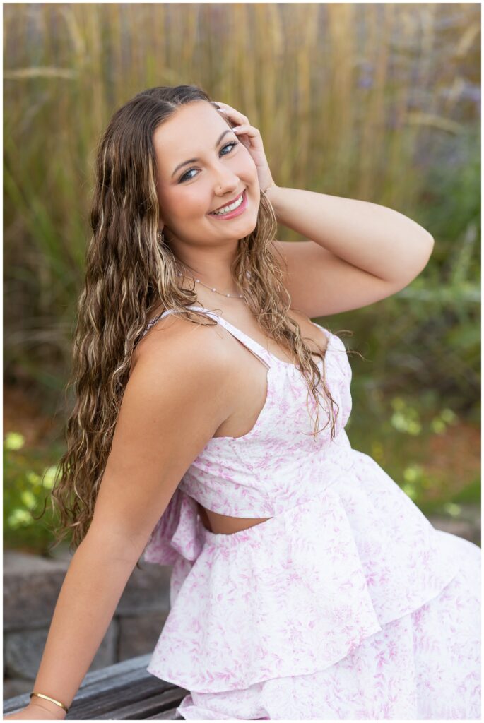 Smiling woman in a light pink floral dress, sitting outdoors on a bench. She has long, wavy hair, and is touching her head. Background is blurred greenery.