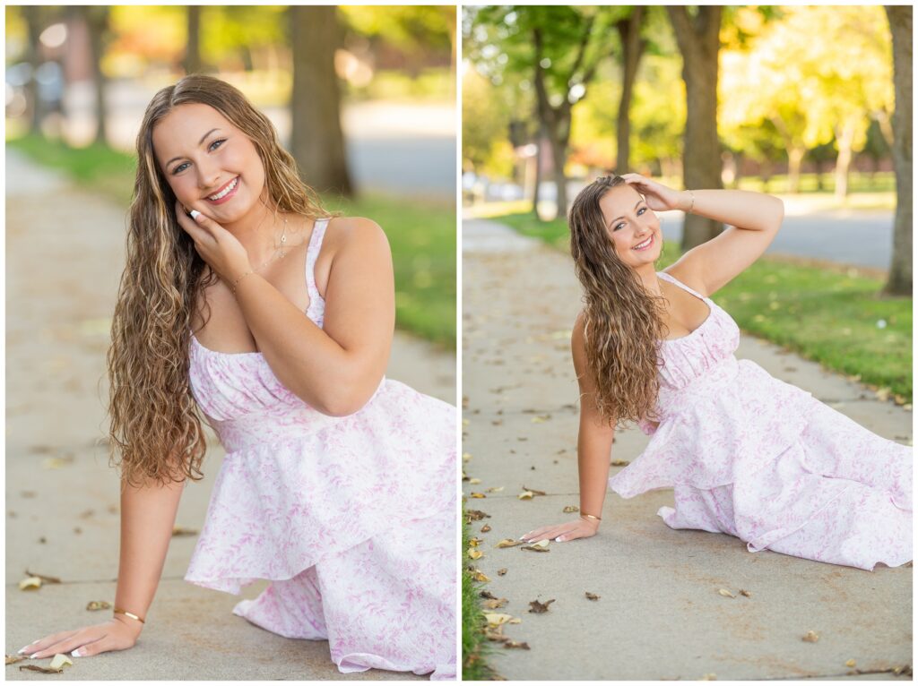 A young woman in a floral dress poses playfully on a sunlit sidewalk. She smiles warmly, with trees and soft-focus greenery in the background.