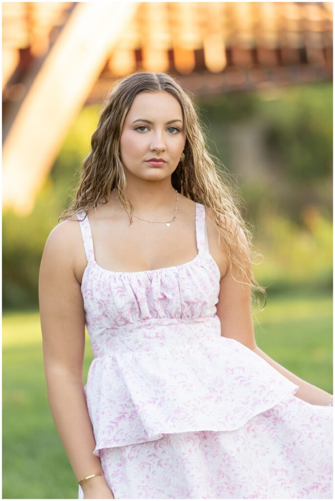 Young woman with wavy hair in a light pink floral dress stands outdoors, looking confident. Sunlight filters through blurred green and wooden background.