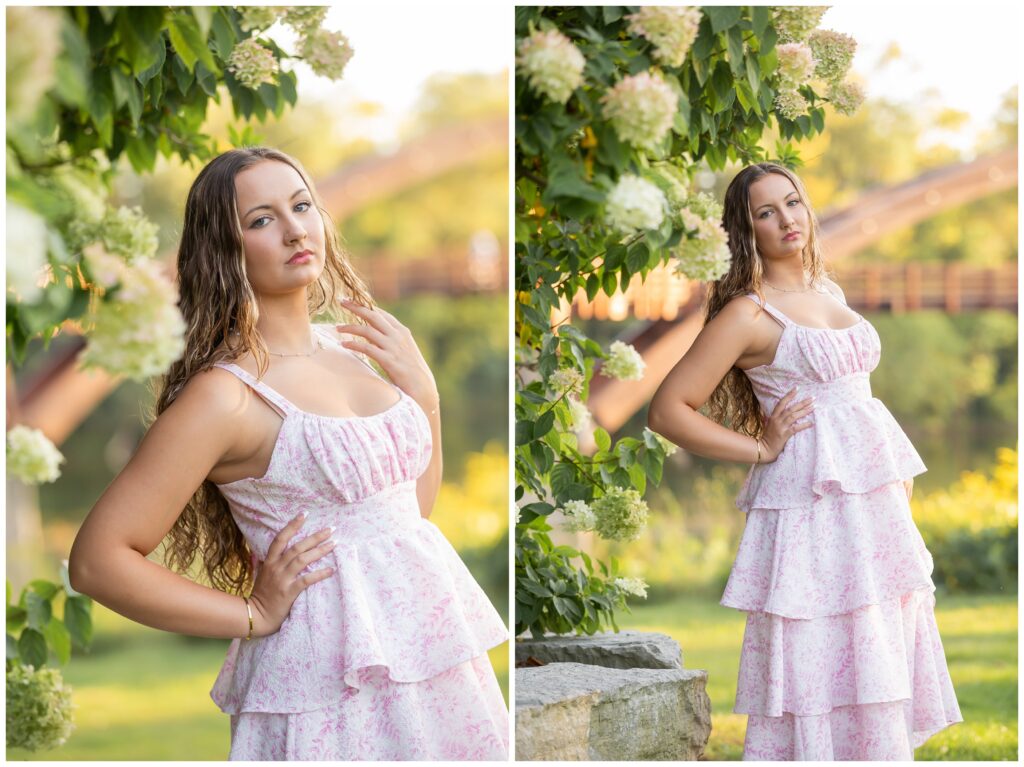 A woman in a pink floral dress poses confidently near blooming hydrangeas. The background features a blurred, sunlit garden and a wooden bridge.