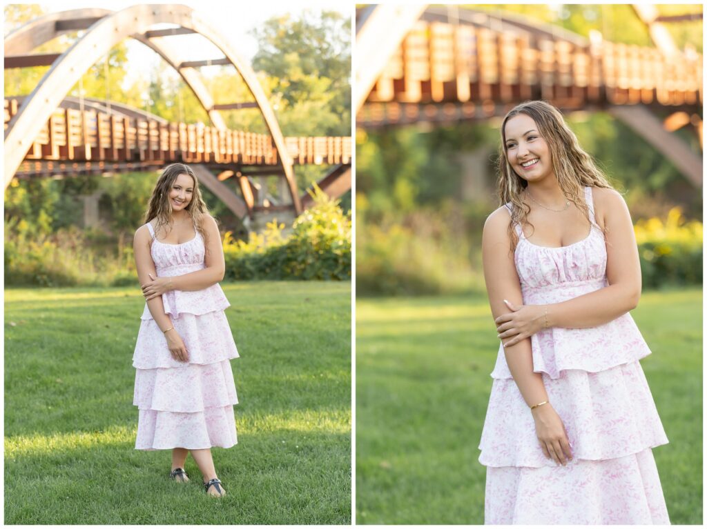 Woman in a tiered pink dress stands on green grass, smiling softly against a backdrop of a wooden bridge and lush trees under sunlight.