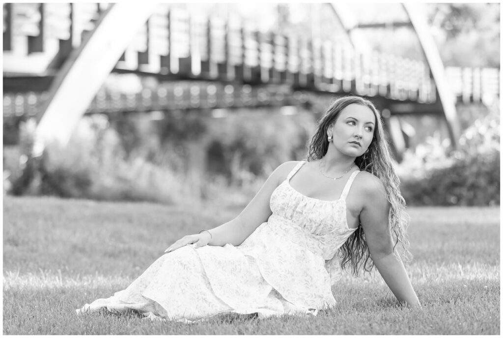 A woman in a white dress sits gracefully on grass, gazing thoughtfully to the side. A blurred bridge arches in the background, suggesting tranquility.
