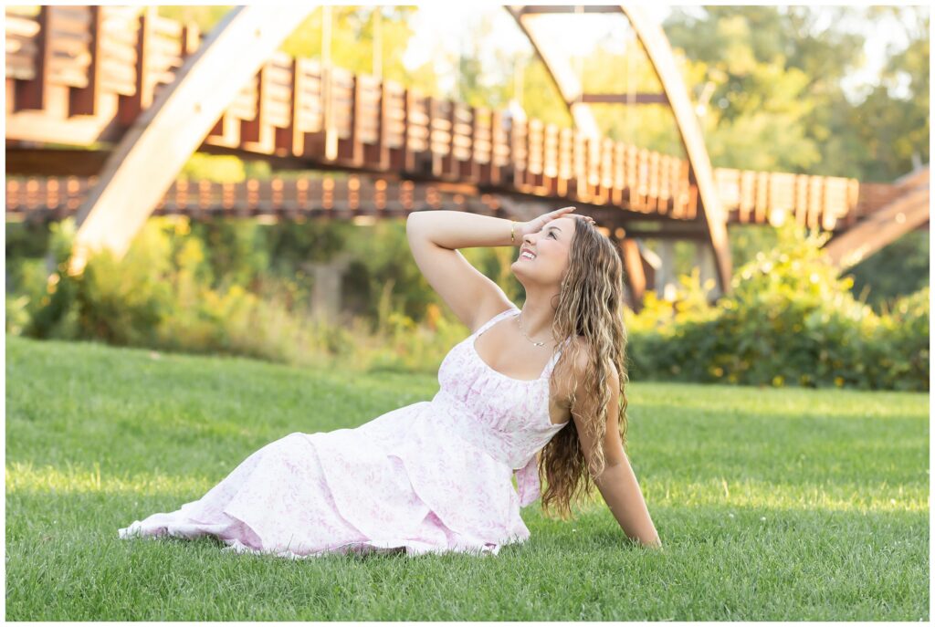 Young woman in a white dress sits on grass, smiling with eyes closed, hand in hair. A wooden bridge and lush greenery are in the background. Serenity.