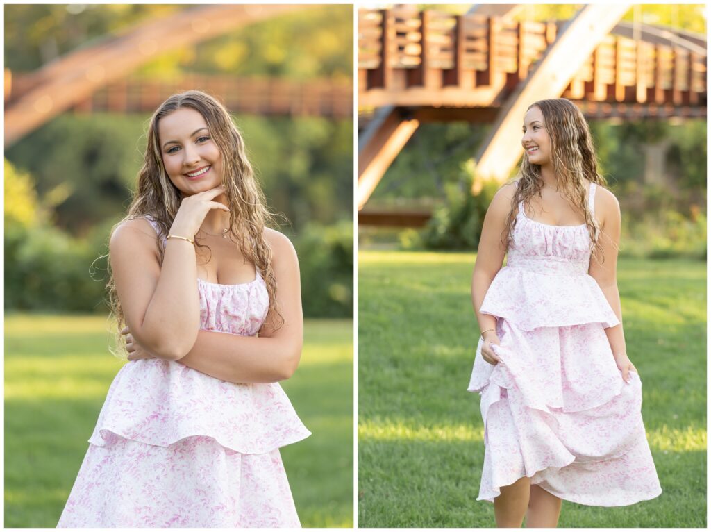 A young woman in a white and pink floral dress poses outdoors with a wooden bridge and greenery in the background, exuding a happy, relaxed vibe.