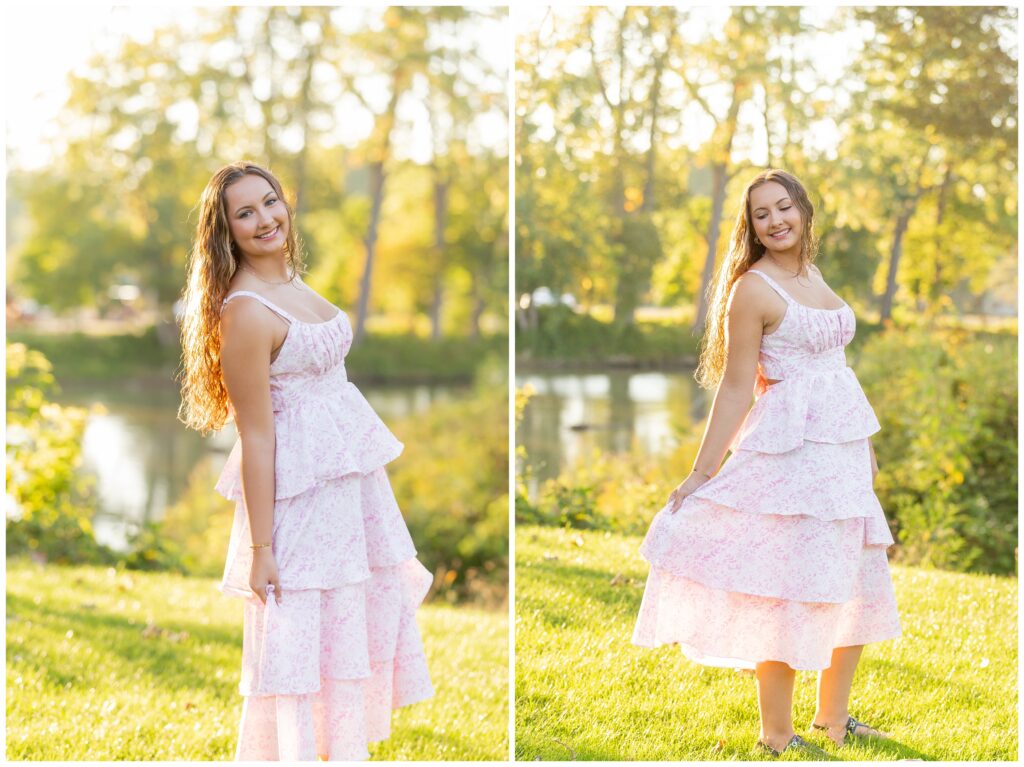 A woman in a light pink, tiered dress stands on grass by a pond, smiling softly. The sun glows through trees, creating a warm, serene atmosphere.
