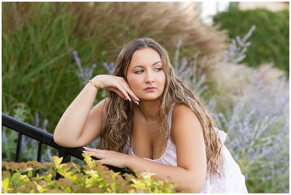 Young woman with long wavy hair leans on a black railing in a garden, surrounded by lush green and purple plants, appearing contemplative and serene.