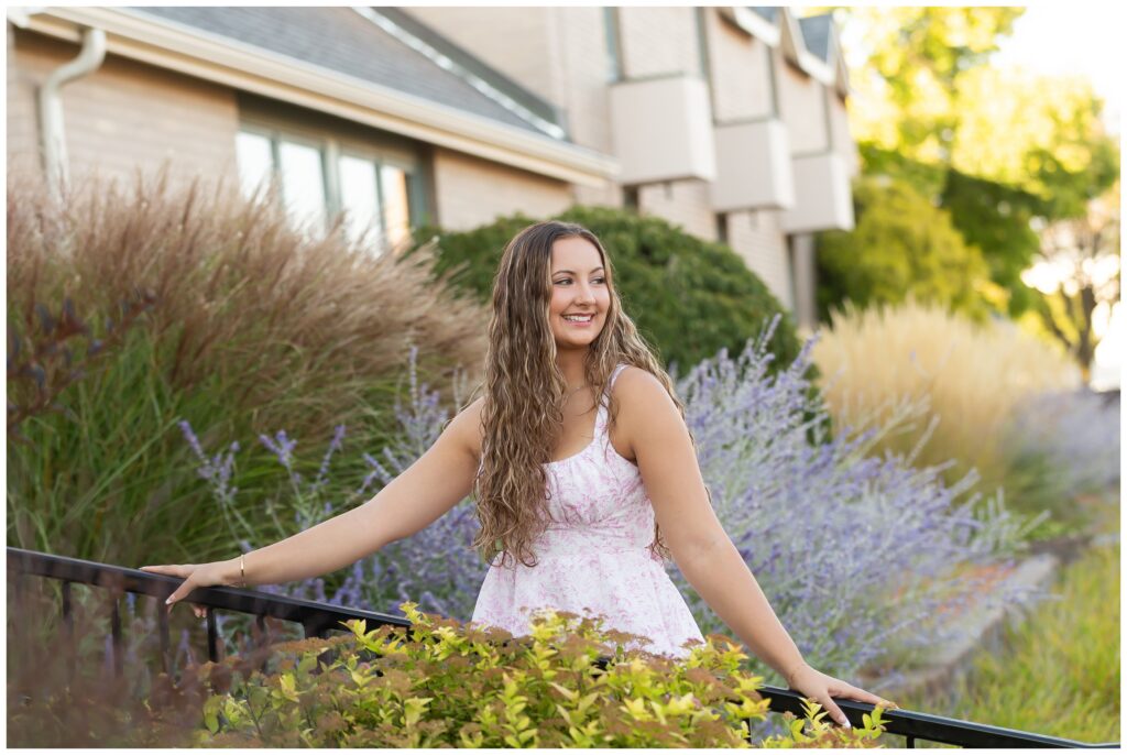 A woman in a floral dress is smiling while standing outdoors, surrounded by lush greenery and lavender plants. The scene conveys a joyful, serene mood.