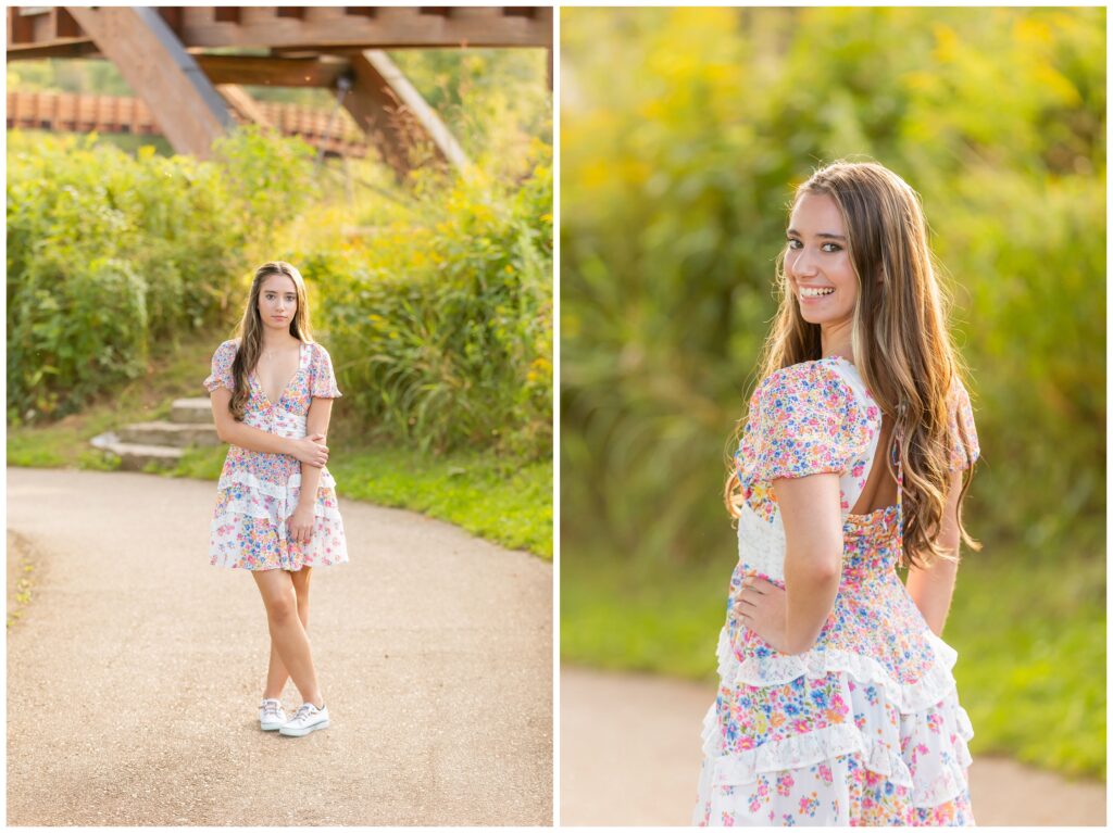 Young woman in a floral dress stands on a park path near greenery, arms crossed, looking serene. Close-up shows her smiling brightly, glowing under soft sunlight.