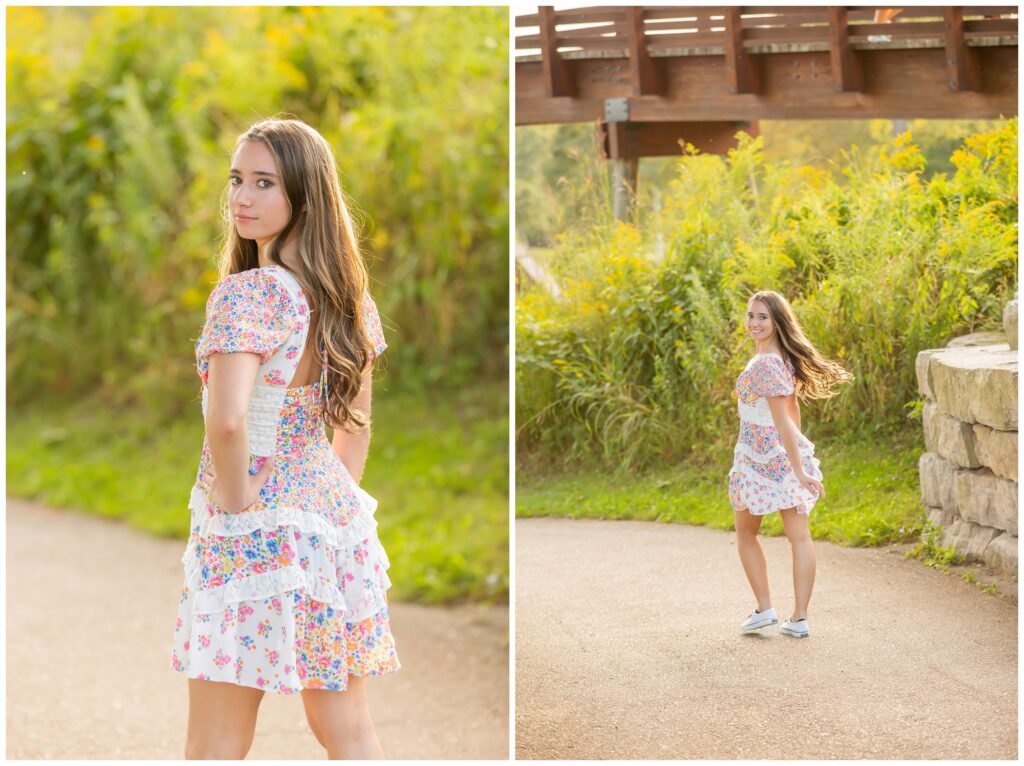 Young woman in floral dress poses outdoors on a sunny day, with greenery and a wooden bridge in the background, conveying a cheerful, natural vibe.