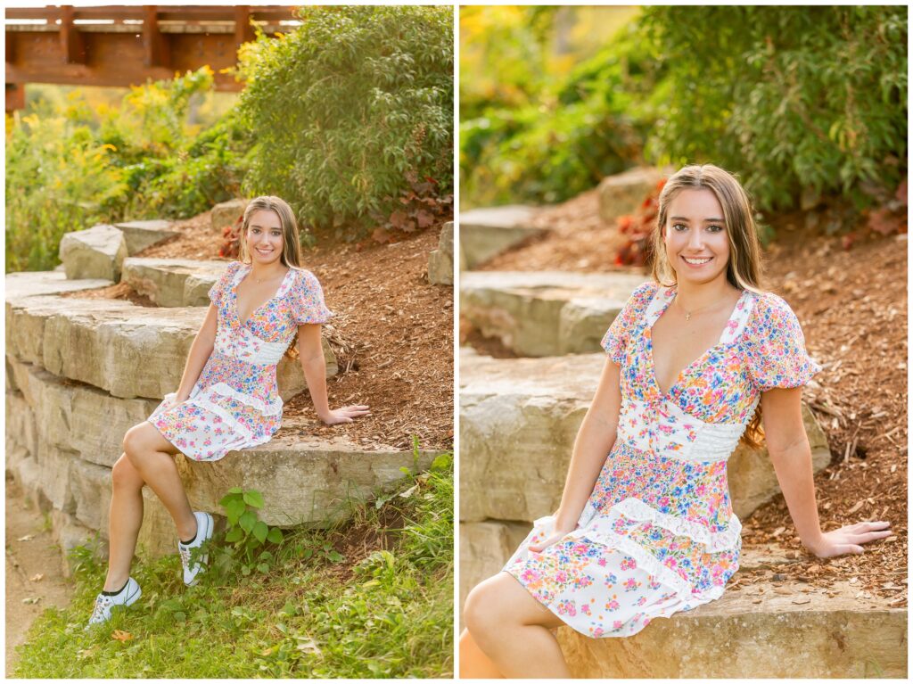 A young woman sits on stone steps in a garden, wearing a floral dress and sneakers. She smiles warmly, surrounded by lush greenery and warm sunlight.