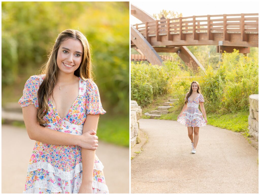 A young woman in a floral dress stands smiling on a sunlit path, with a wooden bridge and lush greenery in the background, creating a warm, cheerful scene.