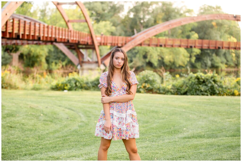 Young woman in a floral dress stands on lush grass, arms crossed, with a wooden arched bridge and greenery in the background, conveying a serene mood.