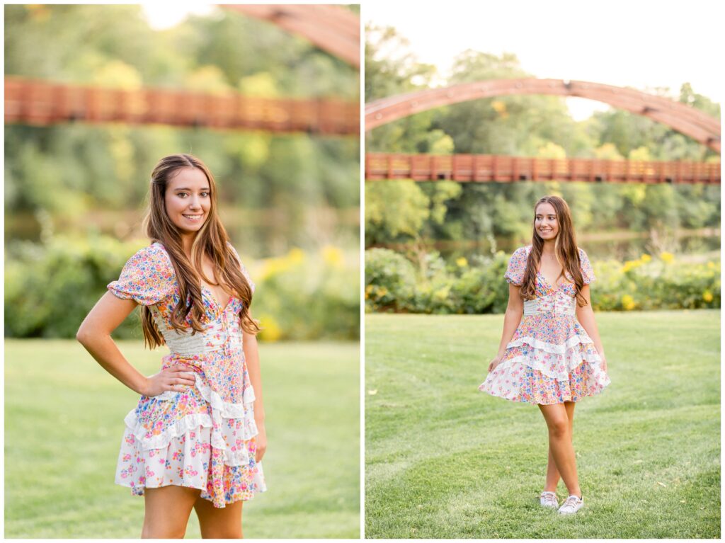 A young woman in a floral dress smiles confidently in a sunlit park. She stands on green grass with a blurred wooden bridge and trees in the background.