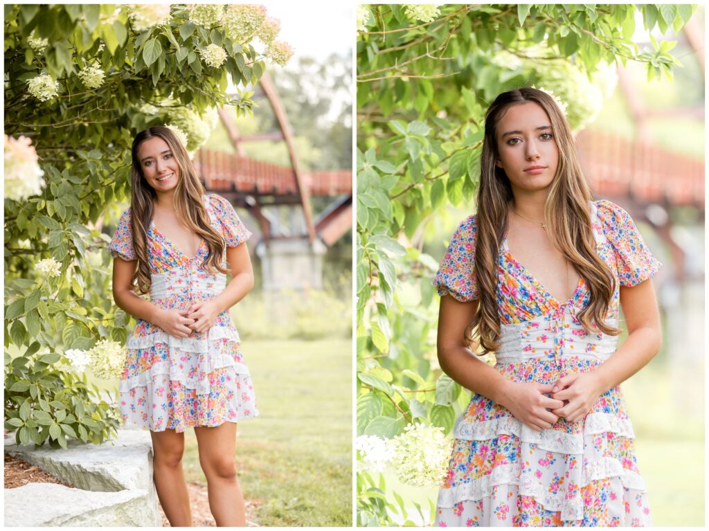 Young woman in a floral dress stands serenely by lush greenery and hydrangeas, with an arched wooden bridge blurred in the background.