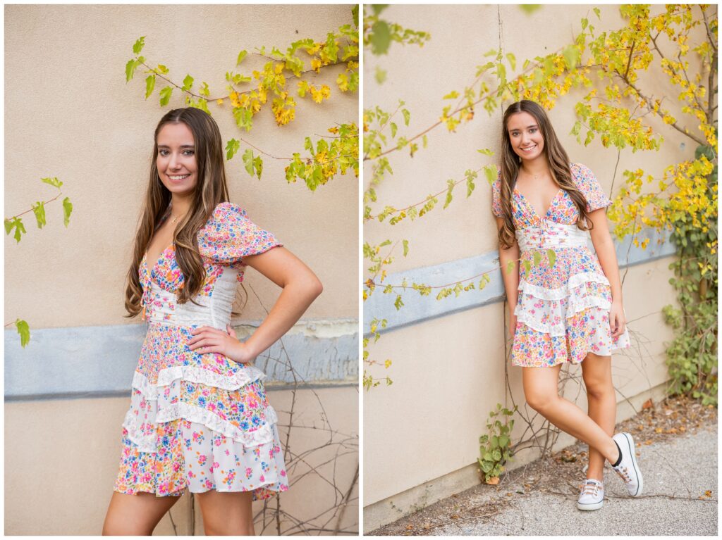 Smiling woman in a floral dress stands against a beige wall with climbing vines. She exudes a cheerful and relaxed vibe, complemented by casual sneakers.