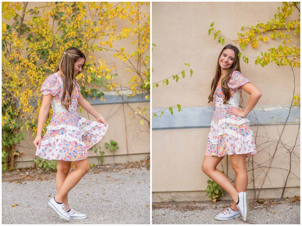 Young woman in a floral summer dress and white sneakers poses joyfully beside yellow-leafed vines against a beige wall, exuding a playful, carefree vibe.
