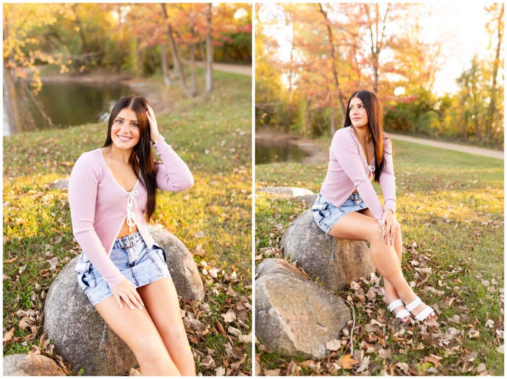 A woman with long dark hair sits on a rock in a park during autumn. She's wearing a pink sweater and denim shorts, smiling warmly. The background features vibrant fall foliage and a calm pond, creating a serene, cheerful atmosphere.