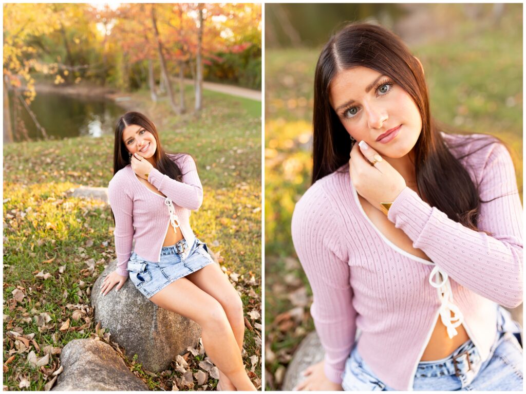 A young woman in a lavender cardigan and denim shorts sits on a rock in a park. Autumn leaves surround her. She looks relaxed and happy.