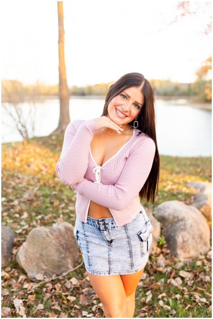 Young woman smiling warmly, standing outdoors on a sunny day by a lake. She's wearing a pink sweater and denim skirt. The scene feels cheerful and serene.