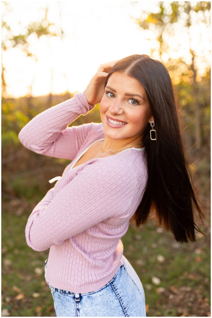 A woman with long dark hair smiles warmly, wearing a pink sweater and jeans. The sun sets behind her, creating a soft, glowing background.